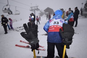 na pierwszym planie rękawiczki narciarskie zawieszone na kijkach narciarskich w tle stok i narciarze